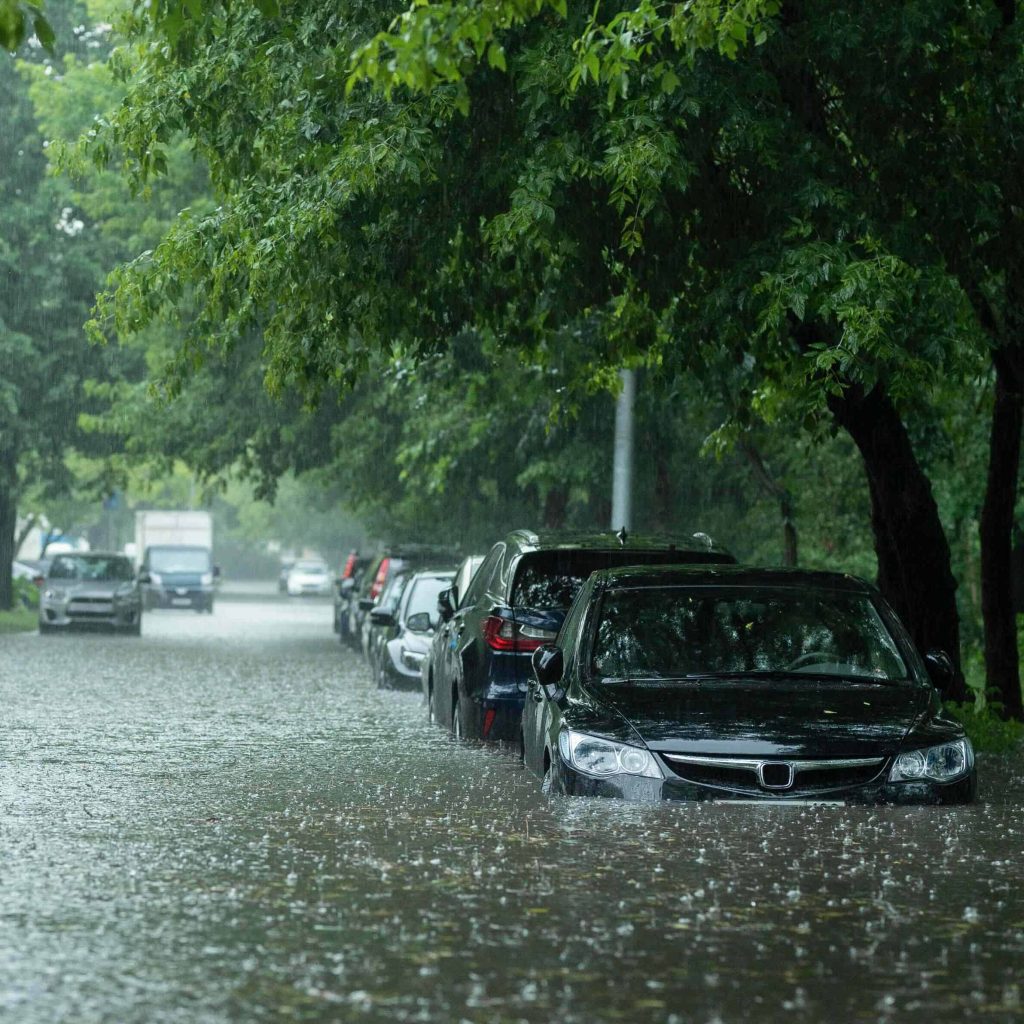 Cars are parked on a street flooded with rainwater, with water reaching up to the lower doors; trees line the street as heavy rain from severe weather continues to fall, creating a gray and gloomy atmosphere.