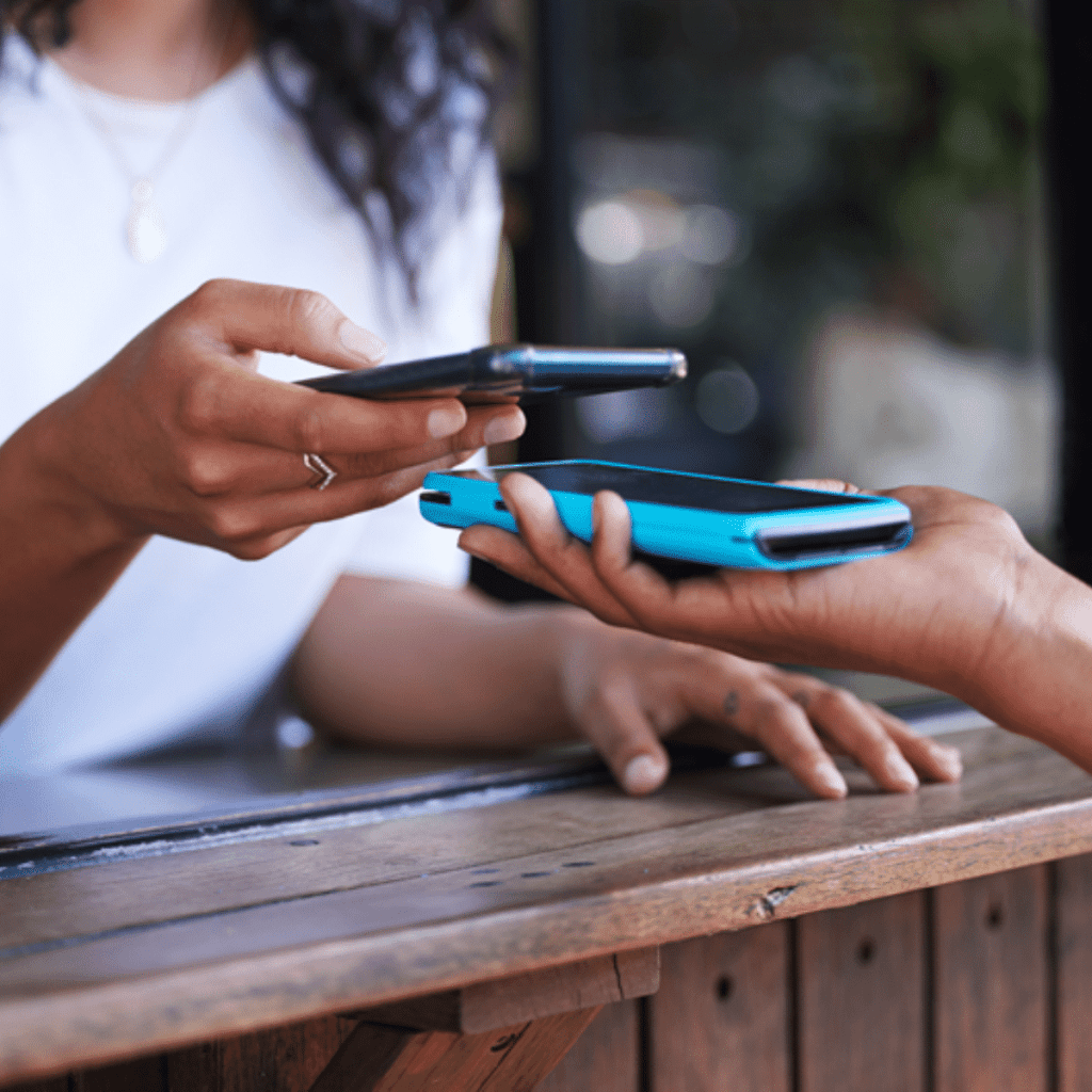 Two people exchange a digital payment by holding their smartphones close together over a wooden counter, highlighting the convenience of contactless transaction technology enabled by mobile banking.