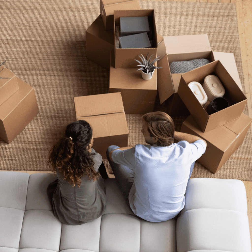 Two homeowners sit on a white couch, seen from above, surrounded by open cardboard boxes with household items inside on a woven rug, suggesting they are moving in or unpacking.