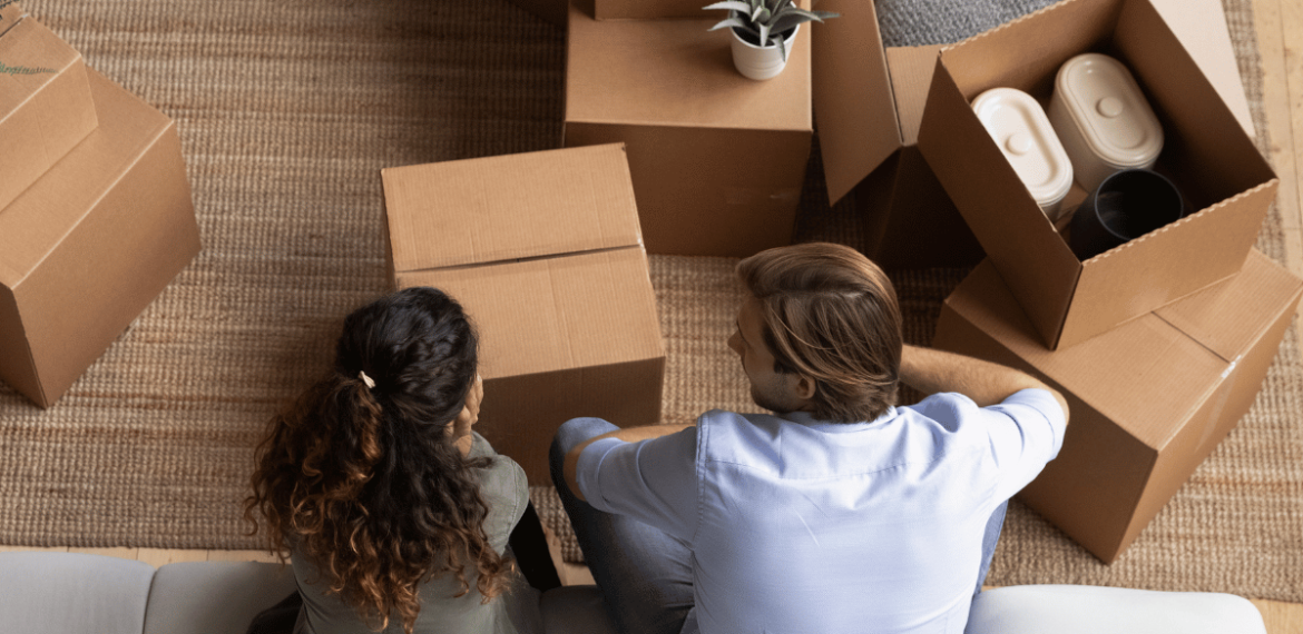 Two homeowners sit on a white couch, seen from above, surrounded by open cardboard boxes with household items inside on a woven rug, suggesting they are moving in or unpacking.