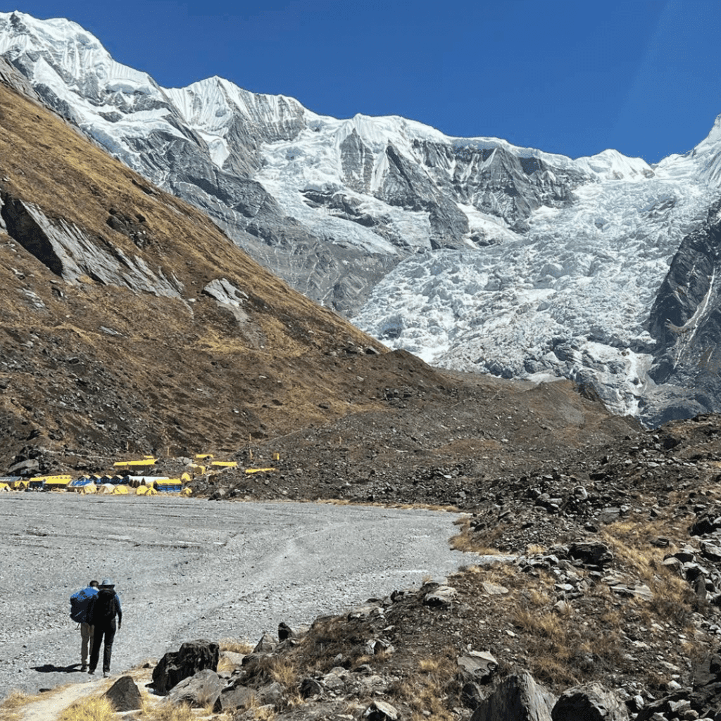 A lone hiker with a backpack walks toward a base camp, marked by a cluster of yellow tents at the foot of snow-covered mountains under a clear blue sky. Rocky terrain and sparse grass cover the ground.