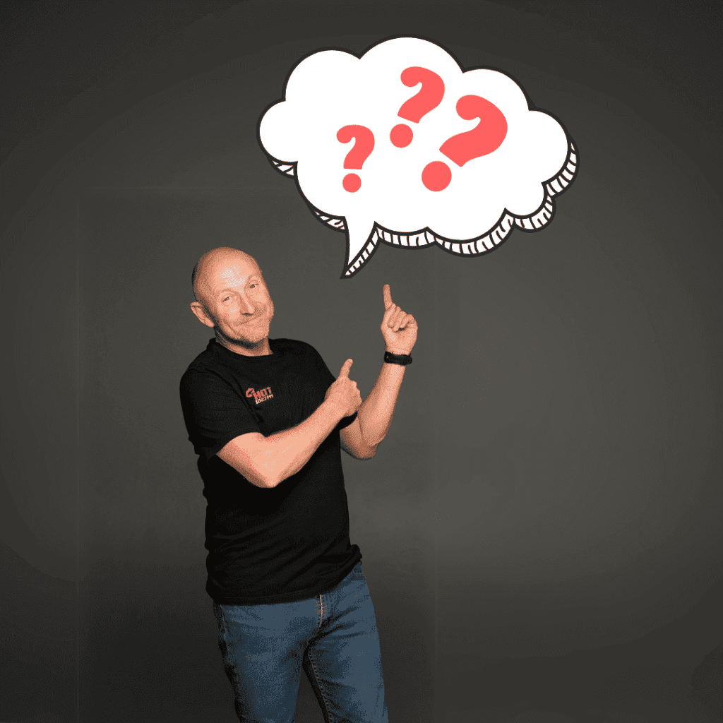 A middle-aged man in a black shirt and jeans stands, smiling and pointing upward at a large illustrated thought bubble filled with red question marks on a dark background.