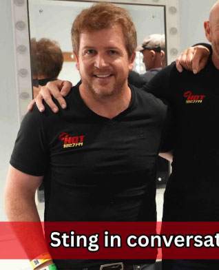 A smiling man with short hair and a beard stands beside another person with an arm around his shoulder. Both wear black t-shirts with red and yellow text. There is a red banner at the bottom with partially visible text reading, Sting in conversat.