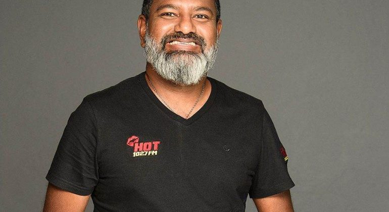 A man with a salt-and-pepper beard and short hair smiles, wearing a black t-shirt with the HOT 102.7 FM logo, standing in front of a plain gray background.