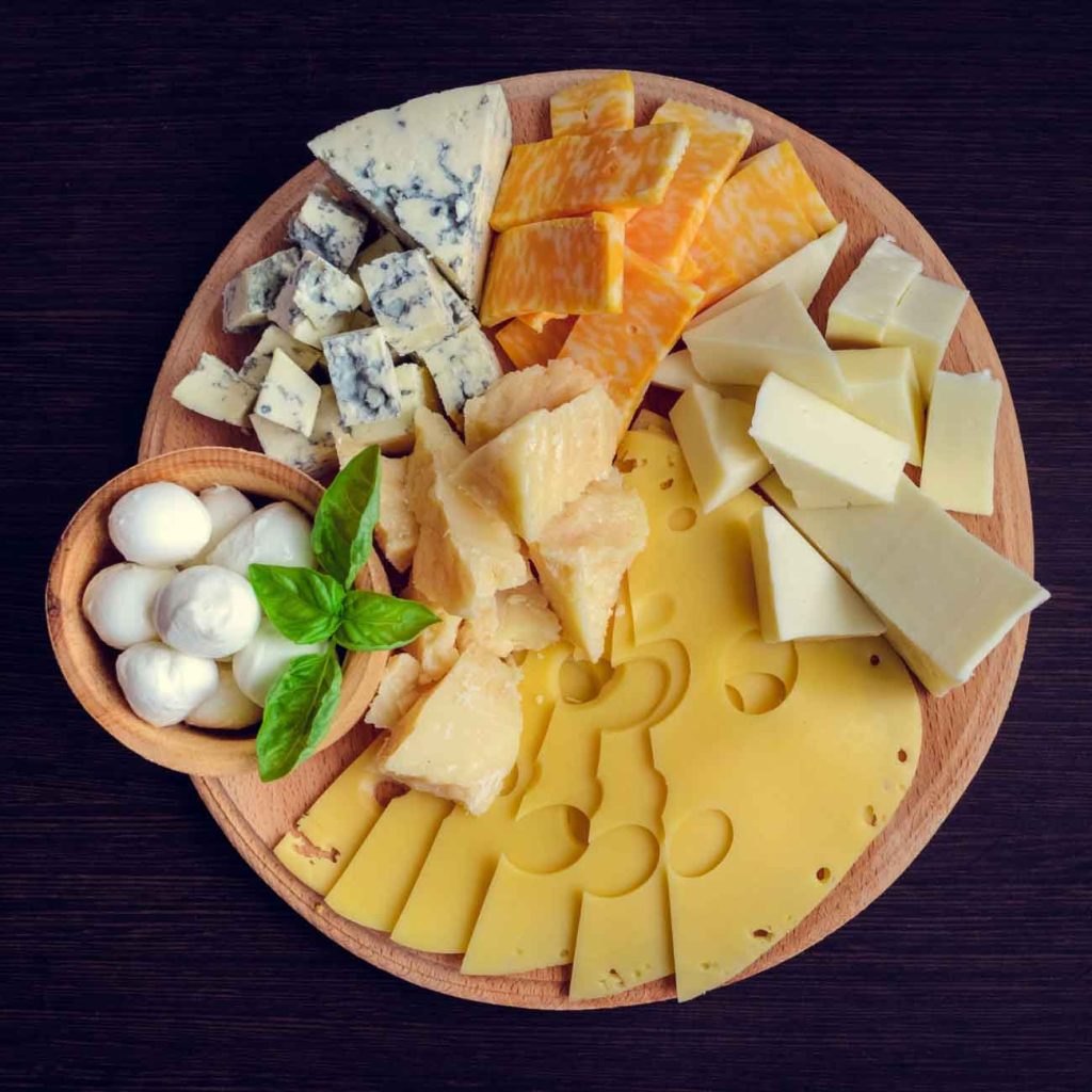 A round wooden board with assorted cheeses, including Swiss, mozzarella balls, blue cheese, cheddar, colby jack, parmesan wedges, and garnished with fresh basil leaves, on a dark surface.