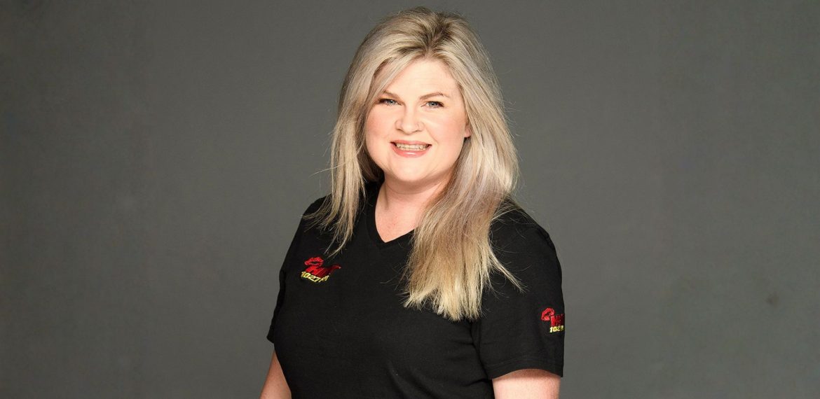 A woman with long blonde hair wearing a black t-shirt stands smiling in front of a plain gray background, celebrating her recent recognition at the New York Festivals Radio Awards.