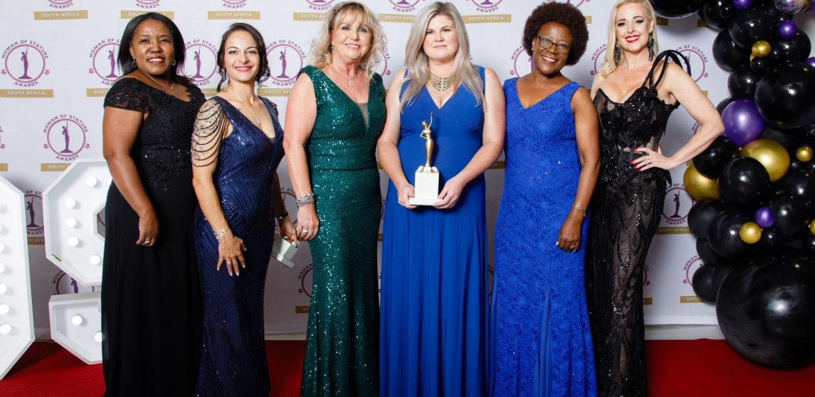 Six women in elegant evening dresses stand on a red carpet at an awards ceremony. The woman in the center holds a trophy, while others smile proudly, with gold and black balloons visible on the right.