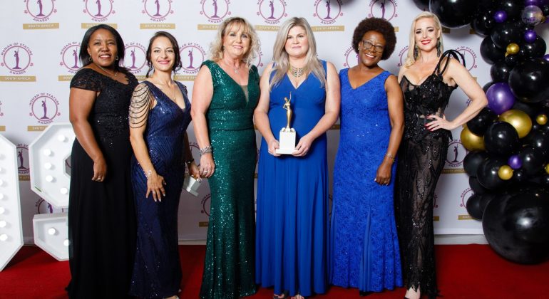 Six women in elegant evening dresses stand on a red carpet at an awards ceremony. The woman in the center holds a trophy, while others smile proudly, with gold and black balloons visible on the right.