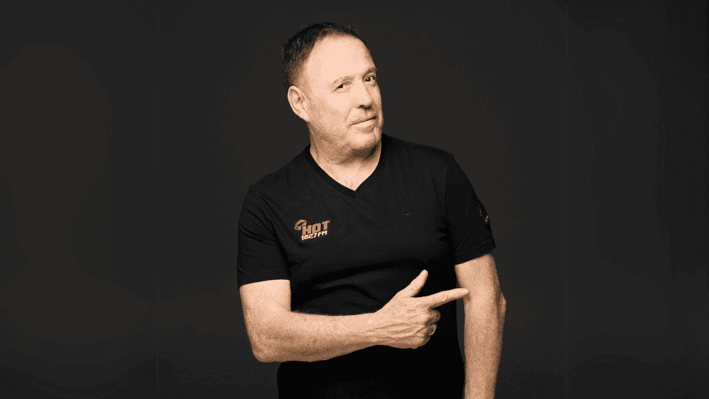 A middle-aged man with short hair wearing a black t-shirt stands against a dark background, smiling slightly and pointing to his left with his right hand.