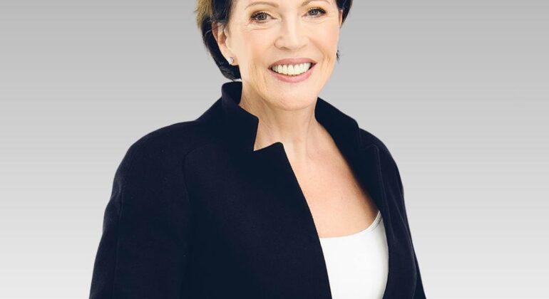 A woman with short dark hair, wearing a black blazer over a white top, exudes hot classic style as she smiles while standing against a plain light gray background.
