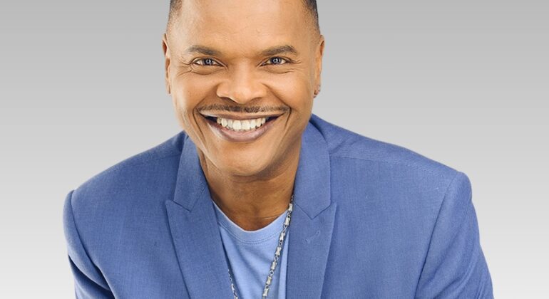 A smiling man with short hair, known as the duke, wearing a light blue blazer, a blue shirt, and a necklace, poses against a gray gradient background.