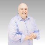 A smiling middle-aged man with short gray hair wears a light blue button-up shirt. He stands in front of a plain gray gradient background, clasping his hands together, ready to discuss the latest hot business trends.