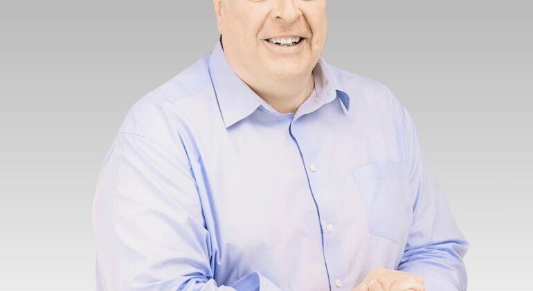 A smiling middle-aged man with short gray hair wears a light blue button-up shirt. He stands in front of a plain gray gradient background, clasping his hands together, ready to discuss the latest hot business trends.