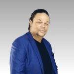 A middle-aged man named Patrick with short curly hair and a goatee wears a blue blazer over a black shirt, standing against a light gray gradient background.