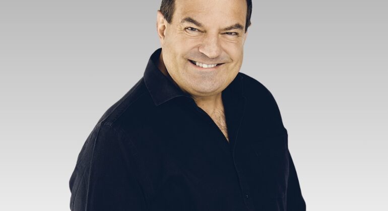 A smiling man with short dark hair wears a black button-up shirt, looking relaxed as if ready for the weekends, standing against a light gray gradient background.