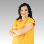 A woman with long dark hair, wearing a yellow short-sleeve blouse, stands with arms crossed and smiles confidently like a sunrise against a plain light gray background.