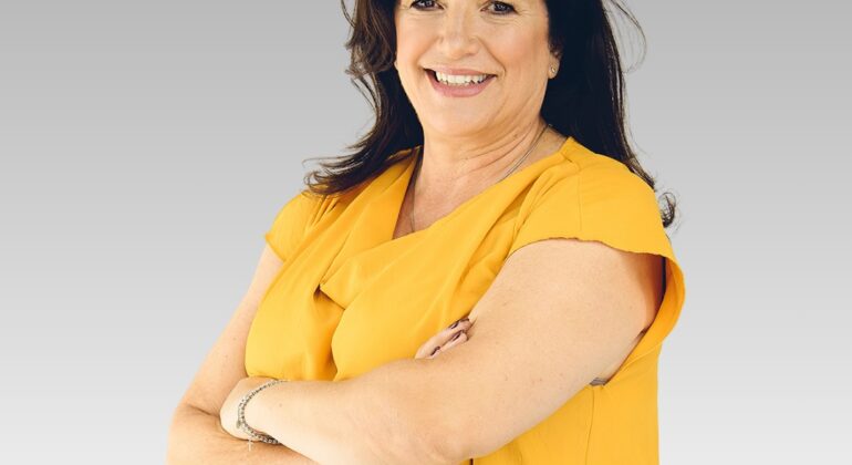 A woman with long dark hair, wearing a yellow short-sleeve blouse, stands with arms crossed and smiles confidently like a sunrise against a plain light gray background.