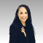 A woman with long, wavy dark hair wearing a black blazer adorned with a decorative brooch, smiling at the camera against a plain light gray gradient background.