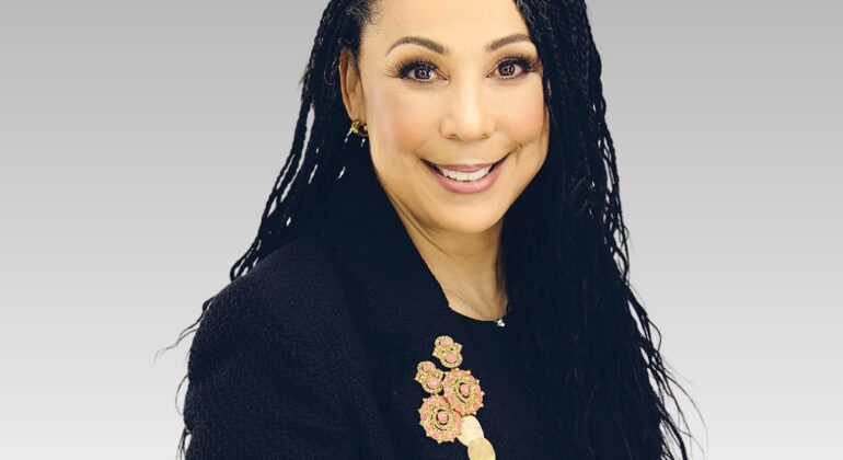 A woman with long, wavy dark hair wearing a black blazer adorned with a decorative brooch, smiling at the camera against a plain light gray gradient background.