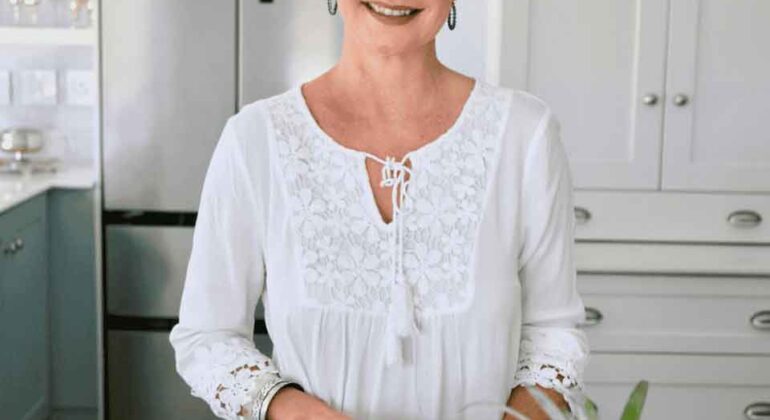 A smiling woman with short blonde hair stands in a bright kitchen, wearing a white blouse and chopping celery on a wooden cutting board with a lemon and pineapple nearby.