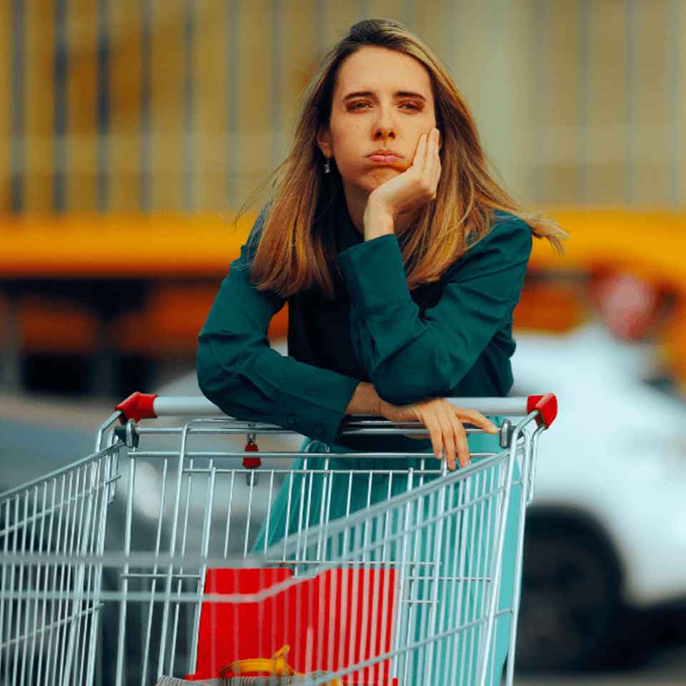 A woman with long blonde hair leans on a shopping cart, resting her head on her hand and looking bored or pensive. She is outdoors, with blurred vehicles and a building in the background.