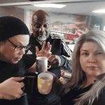 Three adults stand indoors holding mugs, posing for a selfie. One man wears glasses and a beanie, another man in a tracksuit gestures with his hand, and a woman with long, light hair looks at the camera.