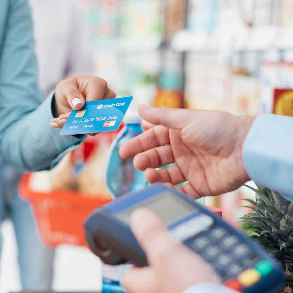 A person hands a blue credit card to another person holding a payment terminal in a grocery store, with a shopping basket filled with groceries visible in the background.