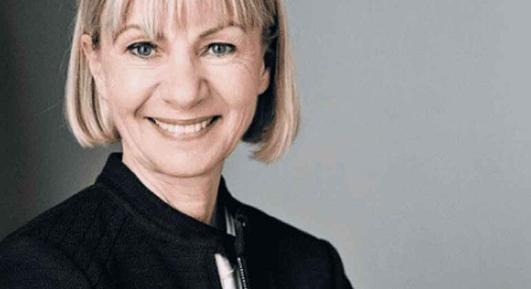 A woman with short blonde hair and bangs is smiling while wearing a dark jacket over a light top, standing against a plain gray background.