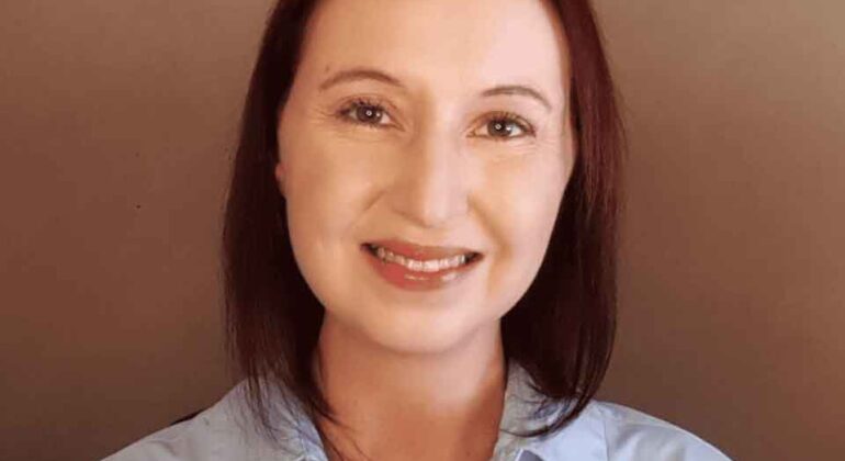 A woman with straight, shoulder-length brown hair smiles at the camera. She is wearing a light blue collared shirt and is posed in front of a plain brown background.
