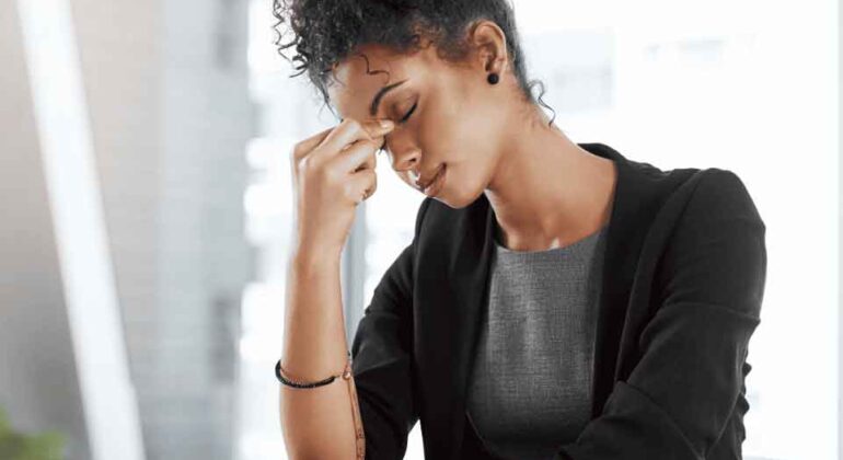 A woman sits at a desk with her eyes closed, pinching the bridge of her nose, appearing stressed or tired. She is dressed in business attire and has a computer and notebook in front of her.