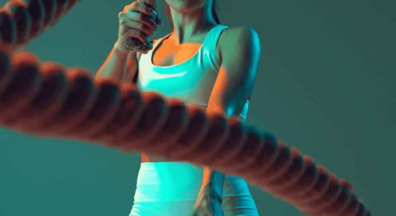 A woman in athletic wear intensely exercises with battle ropes, her face focused and determined, against a teal and orange-lit background.