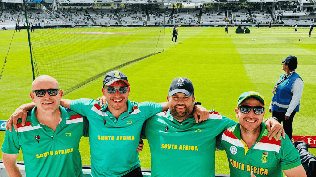 Four smiling men wearing South Africa Proteas cricket shirts pose arm-in-arm at a cricket stadium, with bright green grass, equipment, and other people visible in the background.