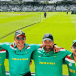 Four smiling men wearing South Africa Proteas cricket shirts pose arm-in-arm at a cricket stadium, with bright green grass, equipment, and other people visible in the background.
