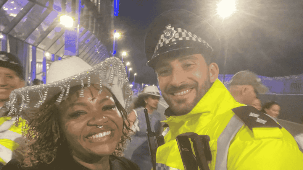 A smiling woman in a white decorative hat, channeling Beyhive energy, stands next to a cheerful police officer in a high-visibility yellow jacket at night, with other people and bright lights in the background.