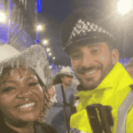 A smiling woman in a white decorative hat, channeling Beyhive energy, stands next to a cheerful police officer in a high-visibility yellow jacket at night, with other people and bright lights in the background.