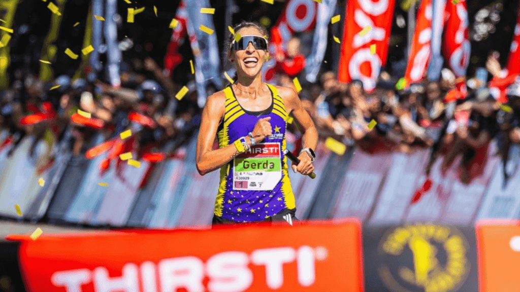 A female runner, Gerda Steyn, smiles as she crosses the finish line, surrounded by cheering crowds and confetti. She wears sunglasses, a purple and yellow athletic outfit, and a race bib reading Gerda.