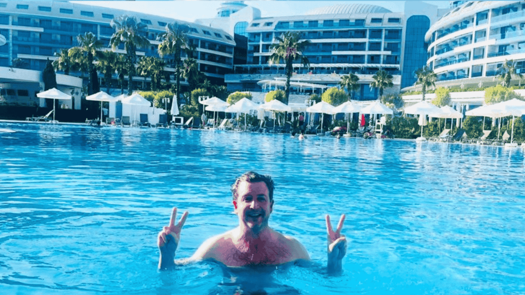 Simon Hill stands in a large outdoor swimming pool in Turkiye, smiling and holding up double peace signs with both hands. In the background are modern resort buildings, palm trees, and white poolside umbrellas.