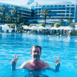 Simon Hill stands in a large outdoor swimming pool in Turkiye, smiling and holding up double peace signs with both hands. In the background are modern resort buildings, palm trees, and white poolside umbrellas.