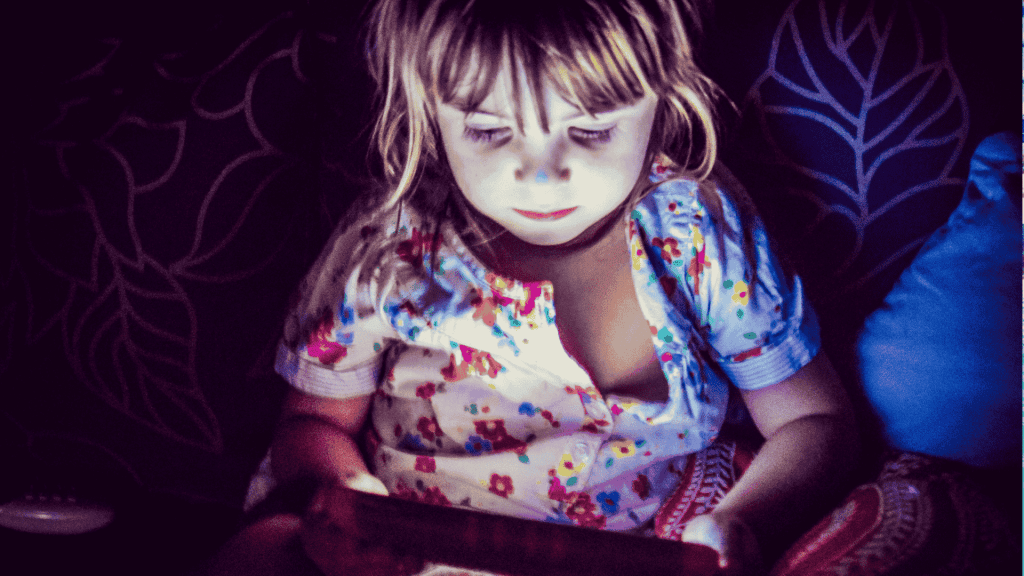 A young child in colorful pajamas sits in dim lighting, illuminated by the glow of a tablet screen, with a patterned cushion and dark leaf-patterned background behind them.