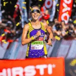 A female runner, Gerda Steyn, smiles as she crosses the finish line, surrounded by cheering crowds and confetti. She wears sunglasses, a purple and yellow athletic outfit, and a race bib reading Gerda.
