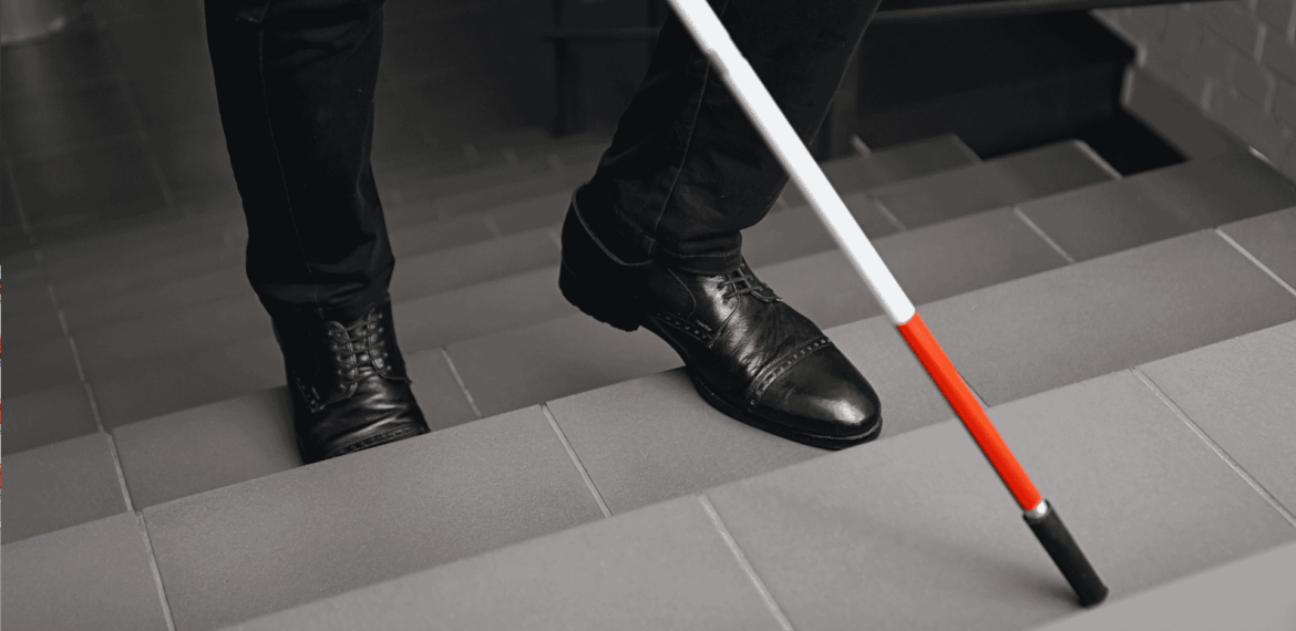 A person wearing black shoes and pants uses a white cane with a red tip to navigate down a tiled staircase.