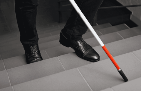 A person wearing black shoes and pants uses a white cane with a red tip to navigate down a tiled staircase.