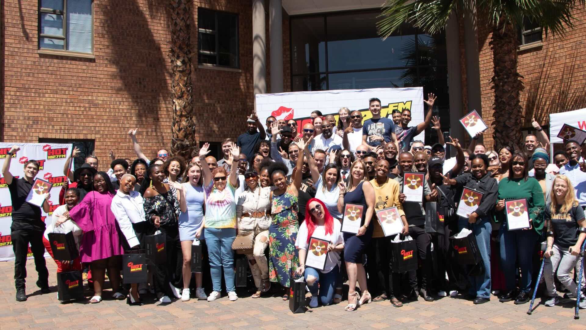 A large group of people stands outside a brick building, smiling, waving, and holding Radio Academy certificates. Some stand on steps, and banners with red and yellow text are displayed in the background.