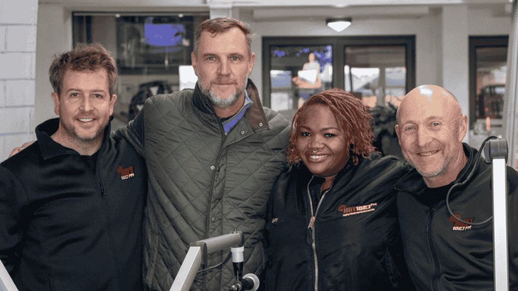 Four people stand closely together, smiling at the camera. Two wear black jackets with Sunshine Tour logos, one sports a green quilted jacket, and another wears a black shirt. Equipment and a TV screen are visible in the background.