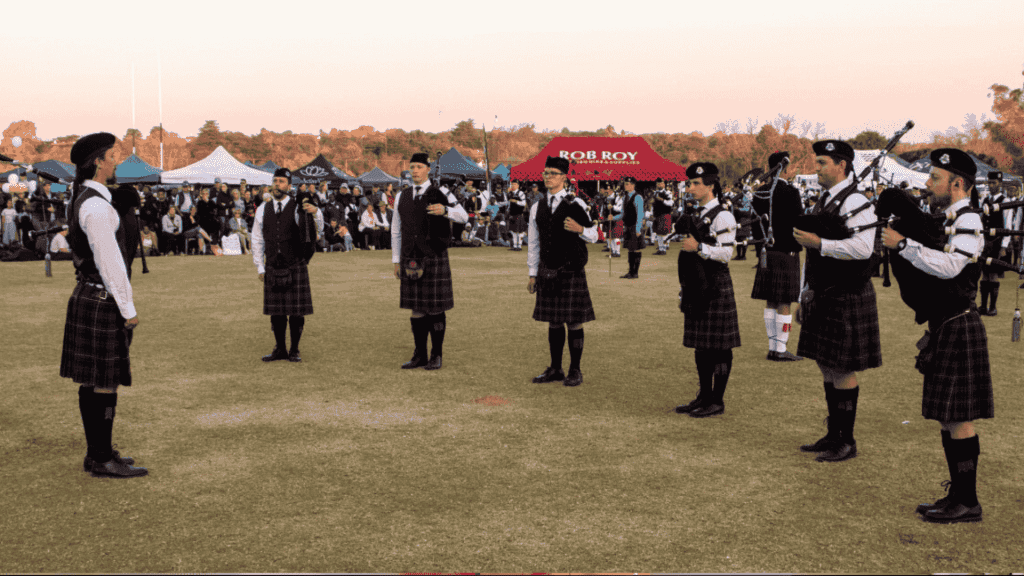 A group of people in kilts and uniforms play bagpipes in a circle on a grassy field during a Highland gathering, with tents and a crowd of onlookers in the background under a clear sky.
