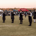 A group of people in kilts and uniforms play bagpipes in a circle on a grassy field during a Highland gathering, with tents and a crowd of onlookers in the background under a clear sky.