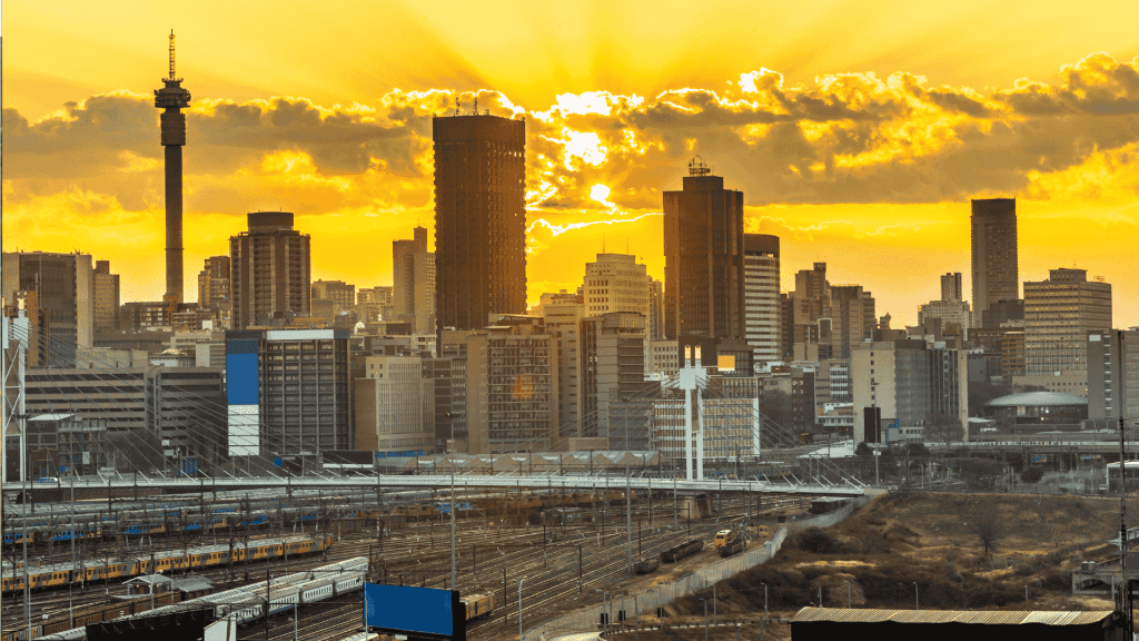 City skyline with tall buildings silhouetted against a dramatic yellow and orange sunset, with train tracks and railway infrastructure in the foreground—a striking view symbolizing efforts to rebuild Johannesburg.