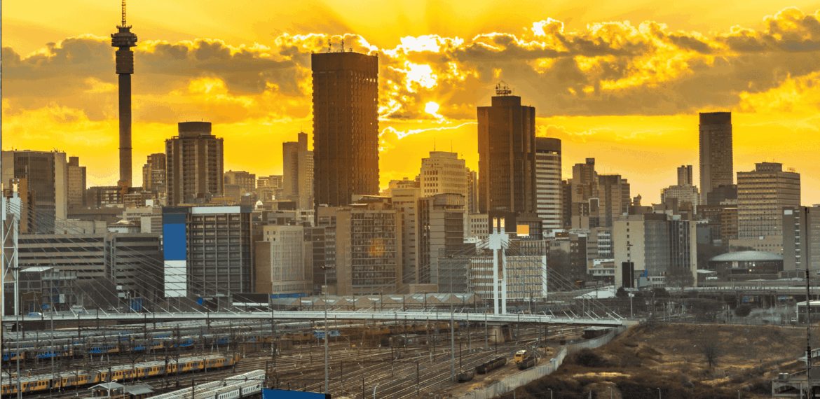 City skyline with tall buildings silhouetted against a dramatic yellow and orange sunset, with train tracks and railway infrastructure in the foreground—a striking view symbolizing efforts to rebuild Johannesburg.