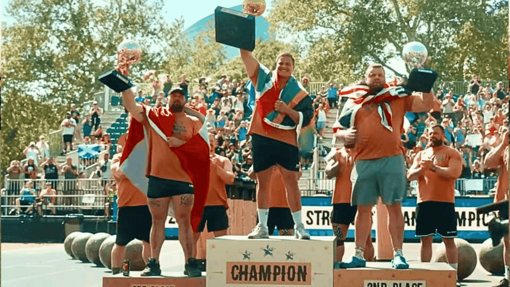 Three men, including Ryno Nel, stand on a winners’ podium at a strongman competition, holding trophies and draped in flags. The central champion raises his trophy high as a cheering crowd and large weights fill the background.
