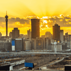 Johannesburg city skyline at sunset with tall buildings, railway tracks, and trains in the foreground under a golden sky—a stunning reminder of efforts to rebuild Johannesburg for a brighter future.